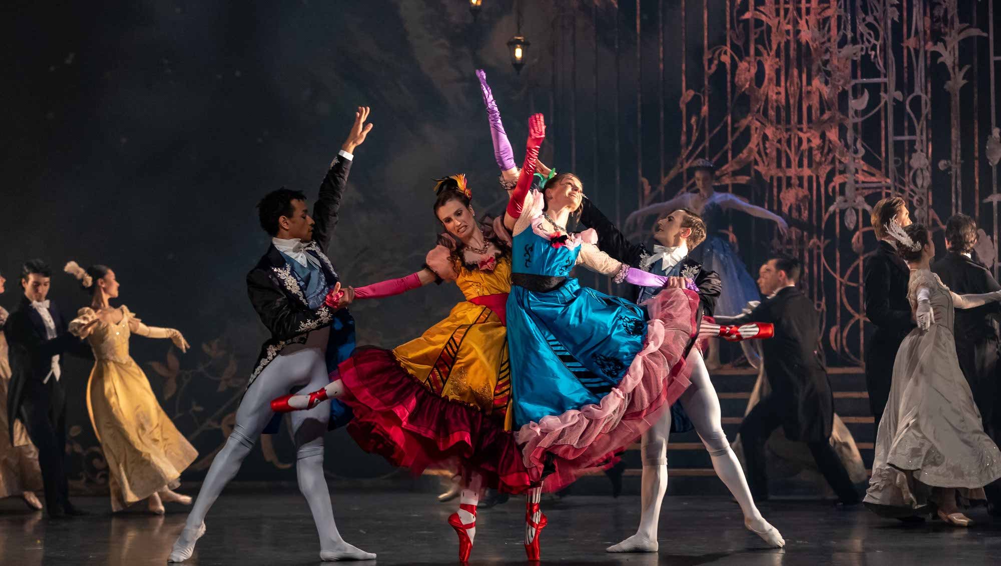 Ballet dancers in colorful costumes perform on stage; two women in bright yellow and blue dresses and red pointe shoes pose playfully with male dancers, with an ornate set and other performers in the background.